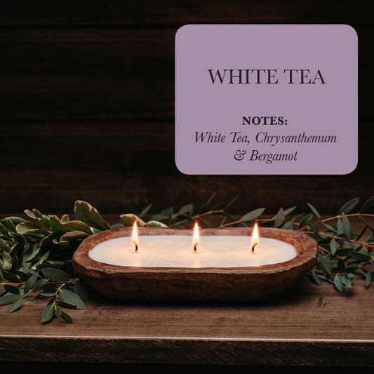 Candle in a wooden holder with greenery and a label for 'White Tea' fragrance on a dark background.